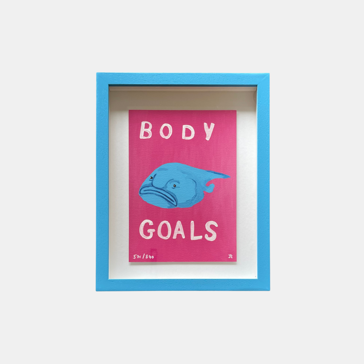 Framed artwork with a pink background, blue fish illustration, and text 'BODY GOALS' on a white wall.